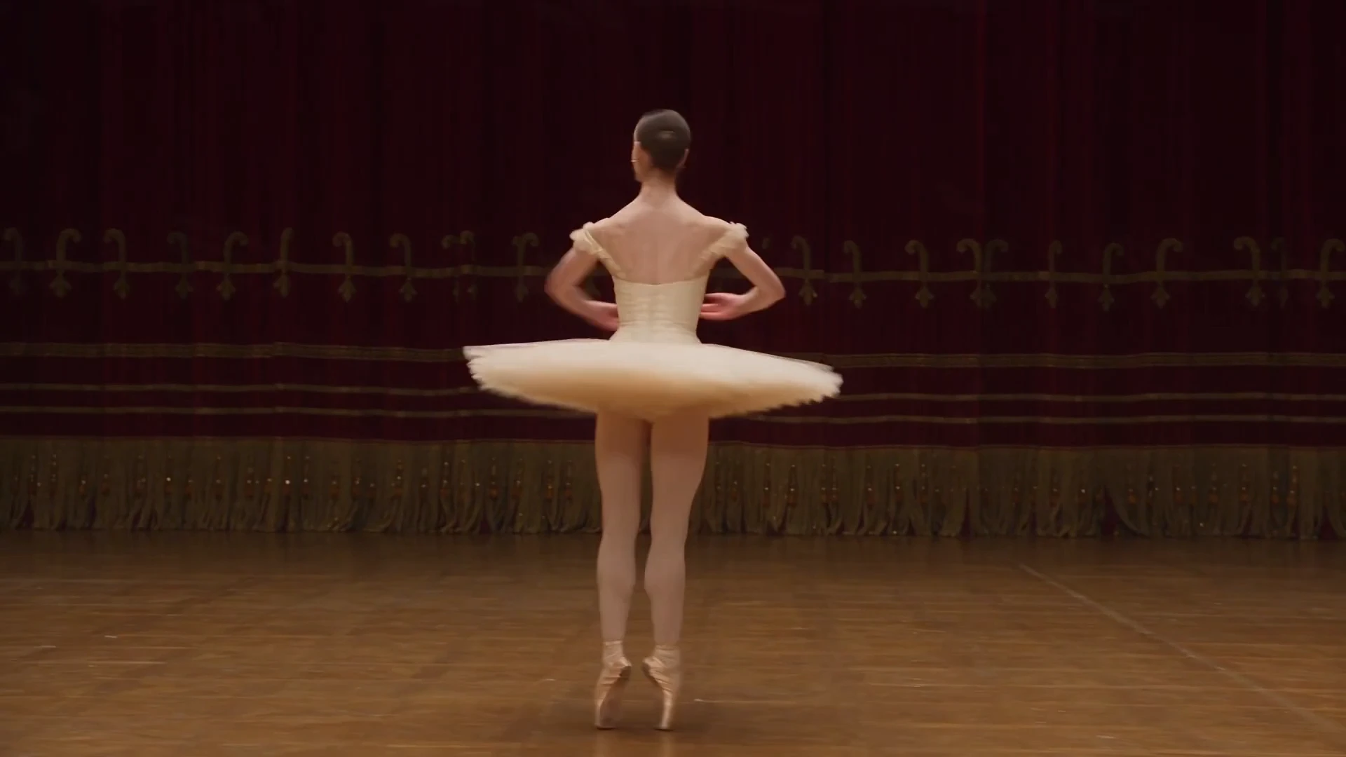 A ballerina in an ivory embroidered costume performing on an ornate opera-house stage.